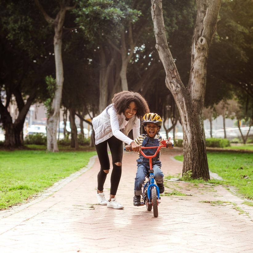 A joyful woman helps a smiling child ride a blue bike on a sunny park path, surrounded by lush green grass and tall trees. The scene conveys happiness and care.
