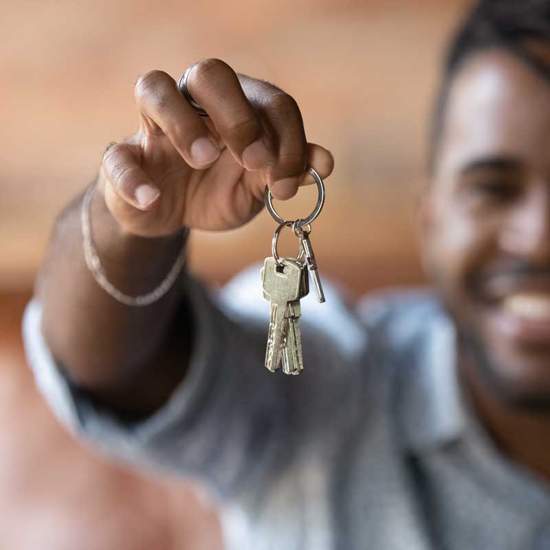 A person holds out a set of keys towards the camera, smiling in the background. The image conveys a sense of excitement and new beginnings.