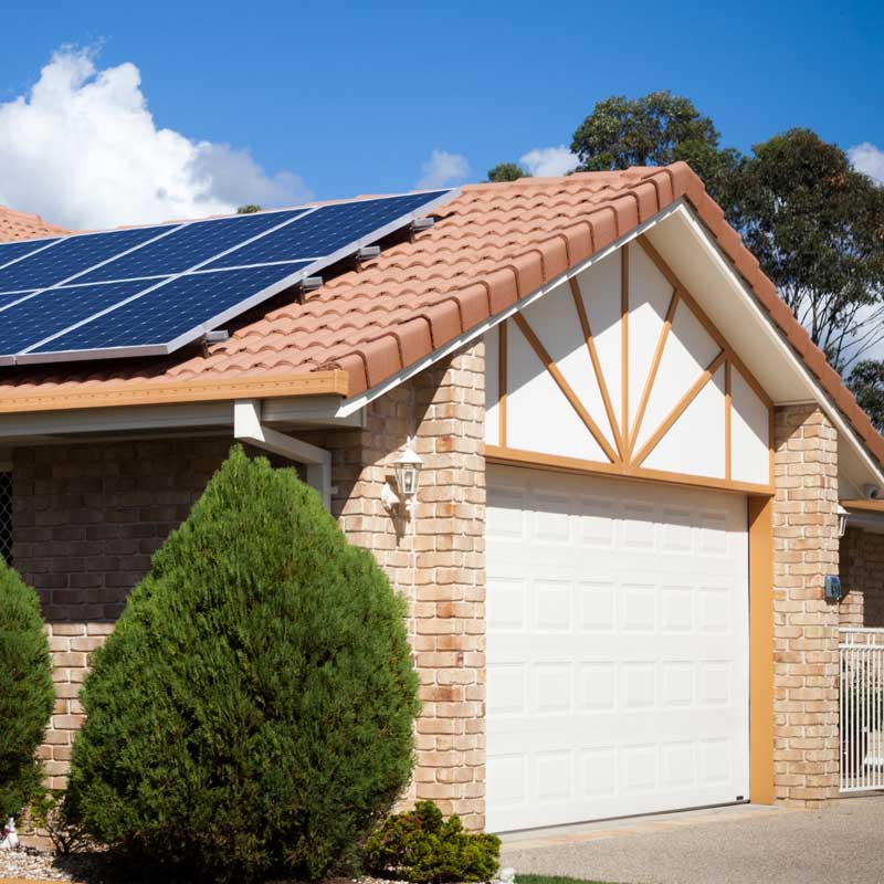 Suburban house with a terracotta roof featuring solar panels, brick facade, white garage door, and green shrubs. Bright, sunny day with blue sky.