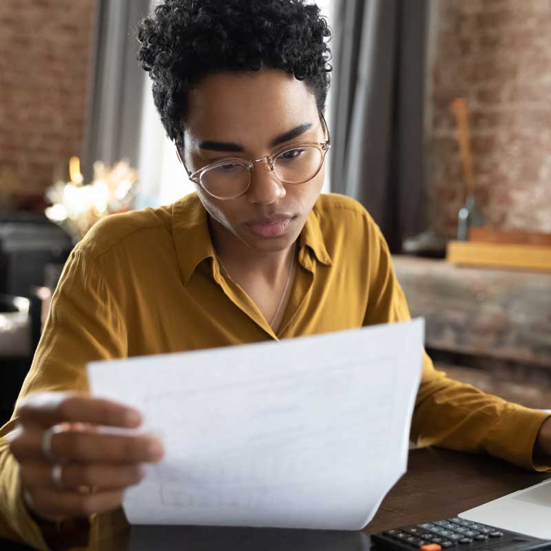Person with glasses in a mustard shirt, focused on reading a document at a wooden desk. Background includes a brick wall and soft lighting, conveying concentration.