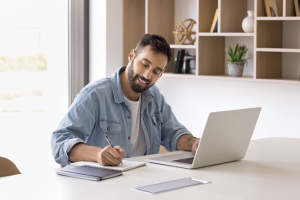 A man is writing on a paper with his laptop on the desk.