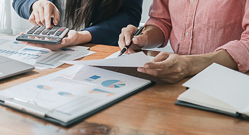 Two people collaborate at a table, using a calculator and analyzing financial charts and graphs. The setting conveys teamwork and focus.