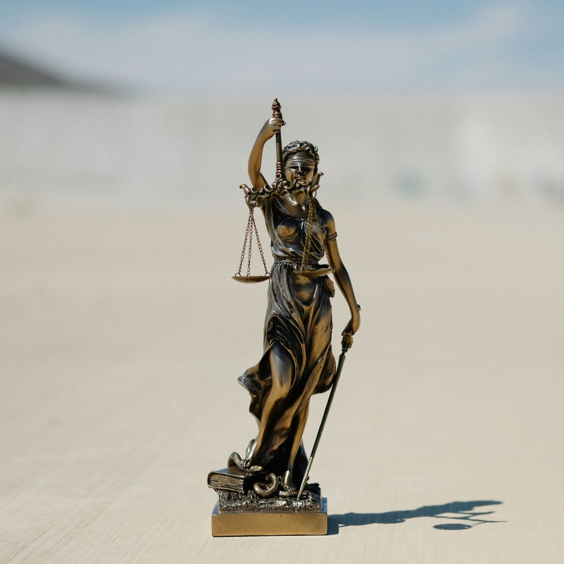 Bronze statue of Lady Justice, blindfolded, holding scales and a sword. The background is a blurred light beige, conveying a sense of fairness.