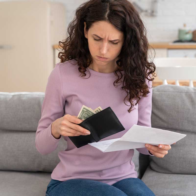 A woman with curly hair wearing a purple sweater sits on a couch, looking concerned. She holds a wallet with cash and a piece of paper, possibly a bill.