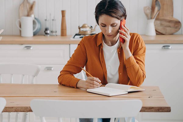 Young woman sits at the kitchen table to make a call while writing notes in a book