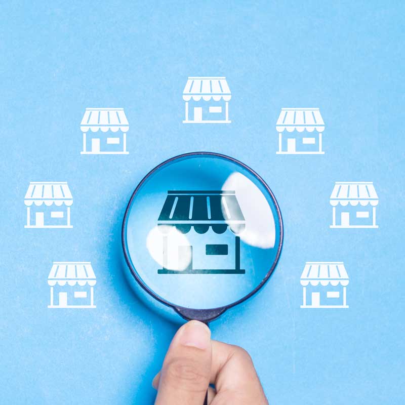 Hand holding magnifying glass over a small shop icon on blue background, surrounded by similar smaller shop icons, symbolizing business focus.