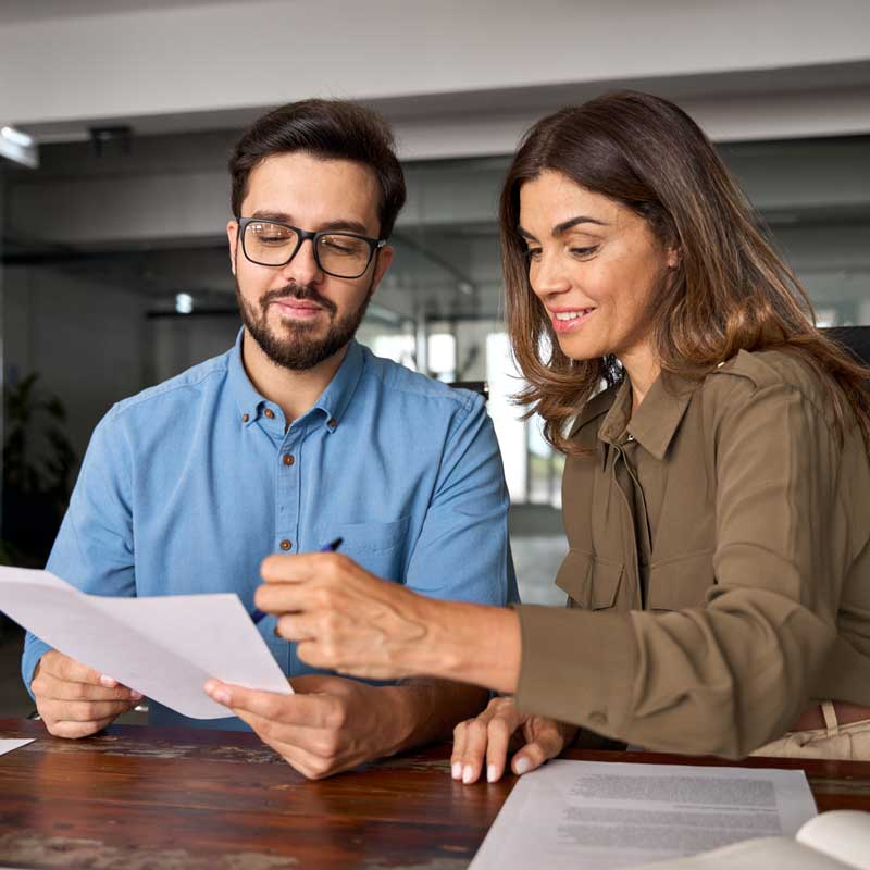 A man in a blue shirt and glasses collaborates with a woman in a brown blouse, both reviewing documents at a wooden desk in a bright office setting.