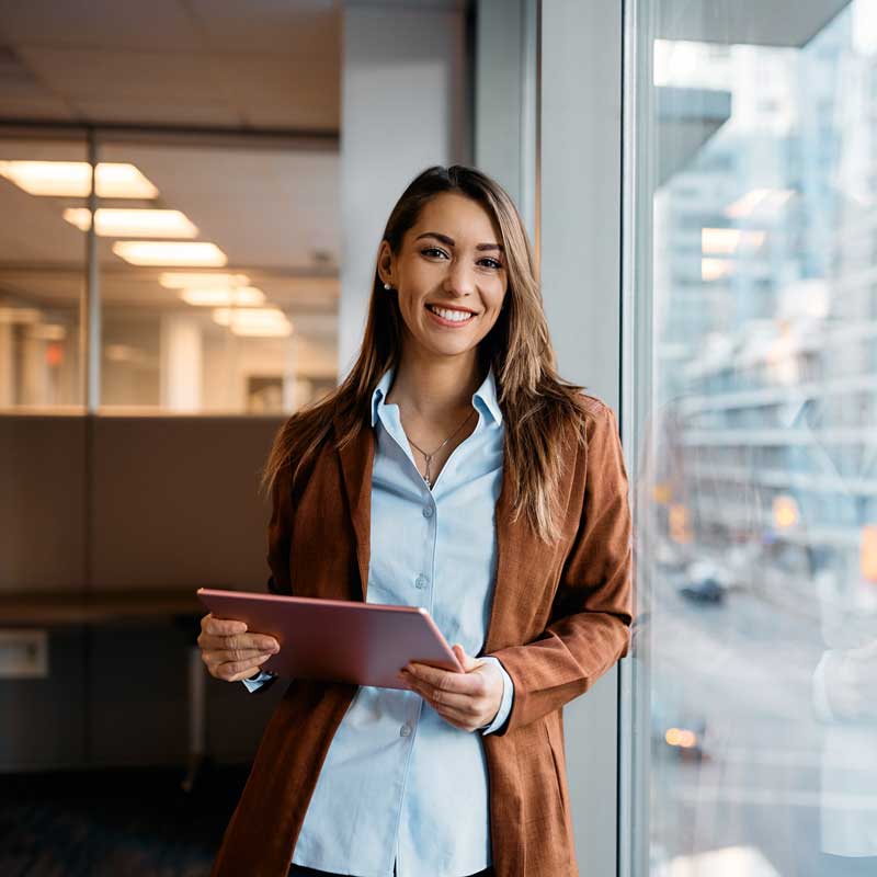 Smiling woman in a brown jacket holds a tablet, standing by an office window. The setting is modern, bright, and conveys a professional tone.