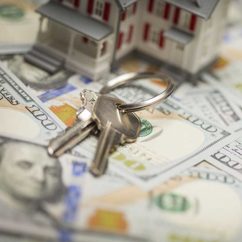 A set of house keys lies on scattered hundred-dollar bills, with a model house in the background, conveying themes of real estate and investment.