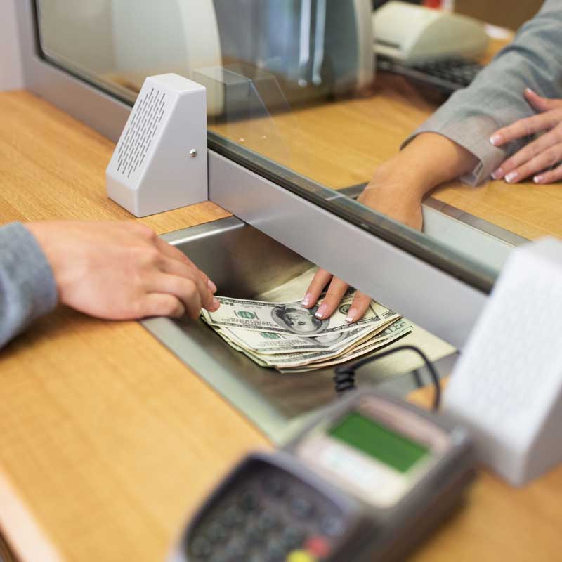 Two hands exchange a stack of hundred-dollar bills through a bank teller window. A credit card terminal is visible, conveying a transactional setting.